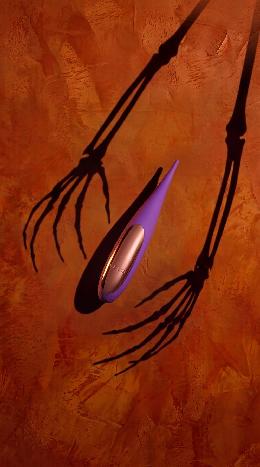 Purple LELO precision massager on an orange plaster wall, held between skeleton-hand shadows