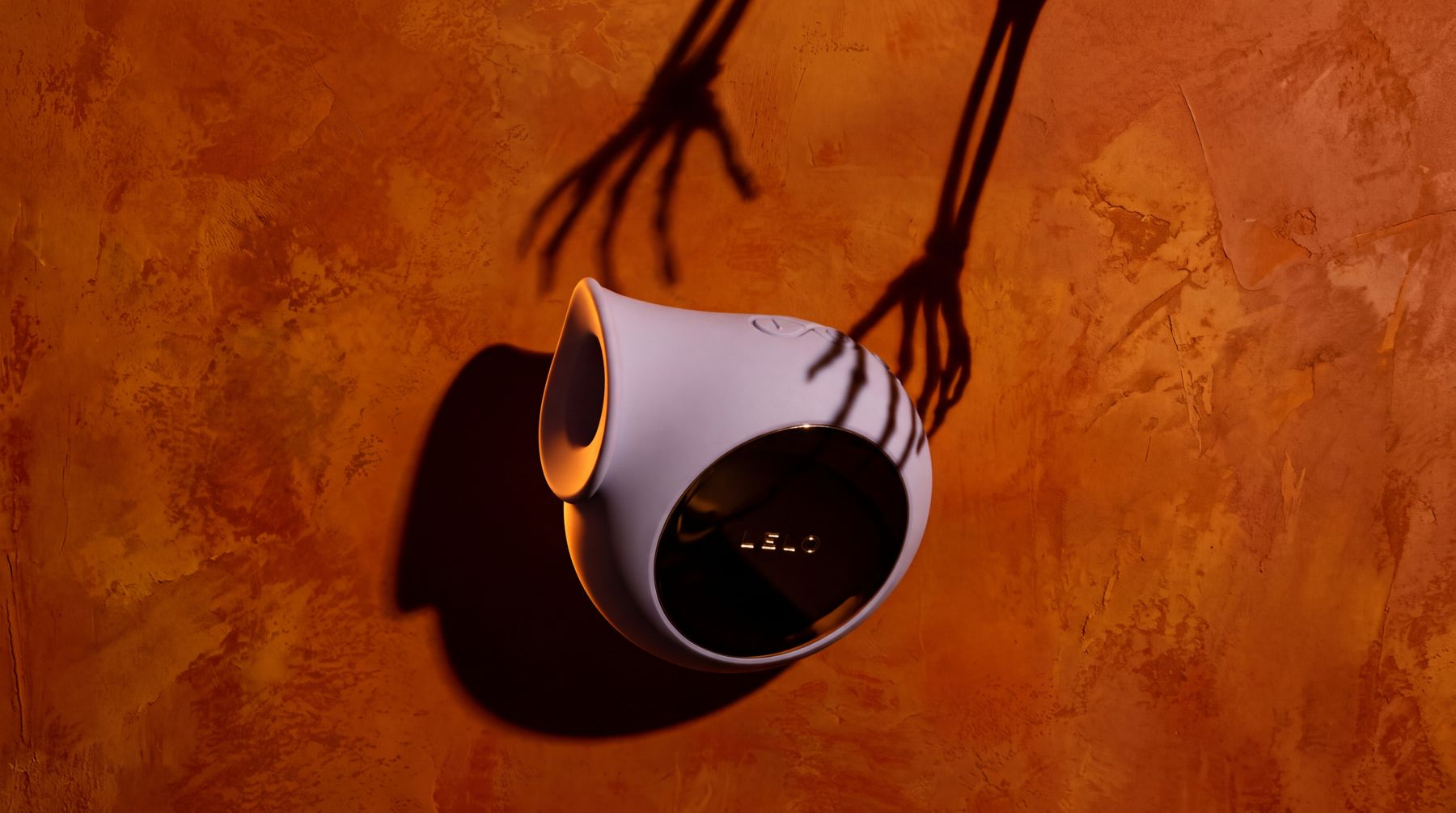 LELO SILA in lilac on an orange plaster wall, with skeleton-hand shadows reaching for it