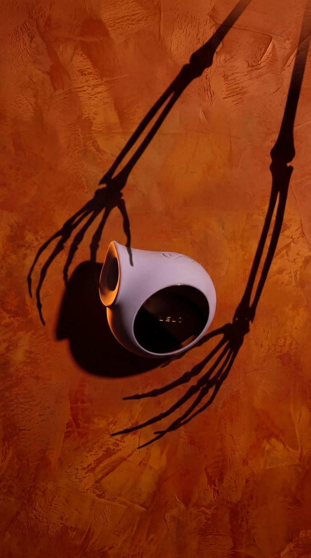 LELO SILA in lilac on an orange plaster wall, with skeleton-hand shadows reaching for it